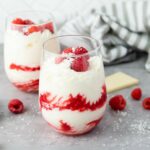Two glass of white chocolate mousse with raspberries filled with a layered raspberry and cream parfait, topped with raspberries and white chocolate shavings.