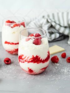 Two glass of white chocolate mousse with raspberries filled with a layered raspberry and cream parfait, topped with raspberries and white chocolate shavings.
