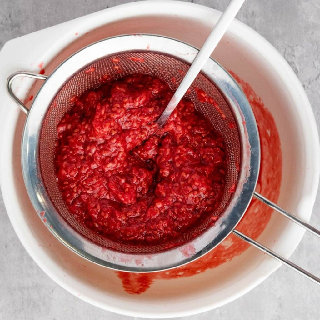 A metal strainer with mashed raspberries sits over a white bowl, with raspberry puree and juice collecting below.