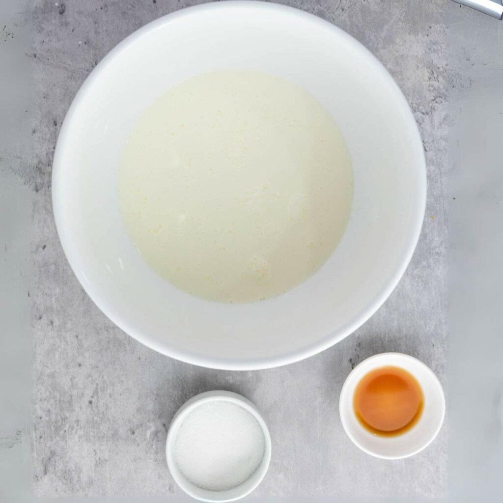 A white bowl with a milky liquid, a small bowl of granulated sugar, and a small bowl of vanilla extract.
