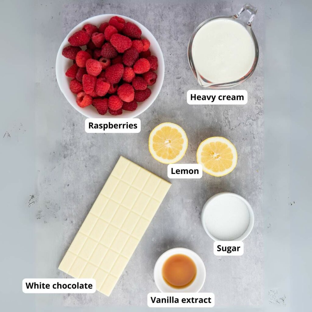 A flat lay of ingredients including raspberries, heavy cream, two lemon halves, sugar, vanilla extract, and a bar of white chocolate.