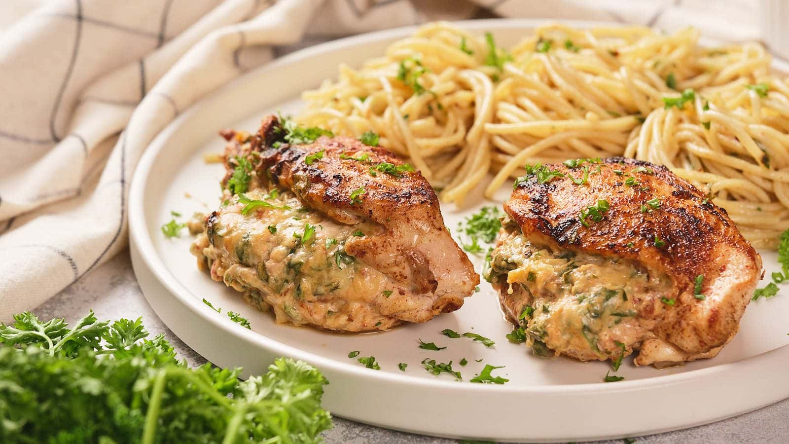 A white plate of air fryer stuffed chicken breast with cooked pasta, and a fresh parsley next to it on a white surface.