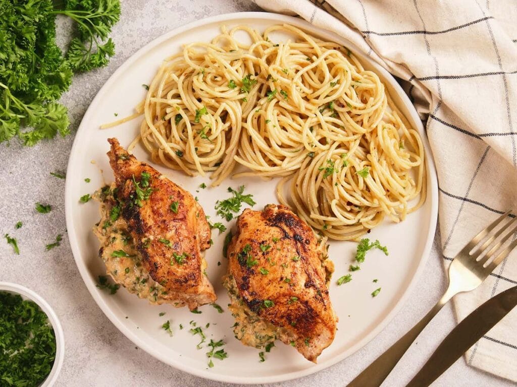 A white plate of air fryer stuffed chicken breast next to fork and knife, garnished with parsley on a light surface.