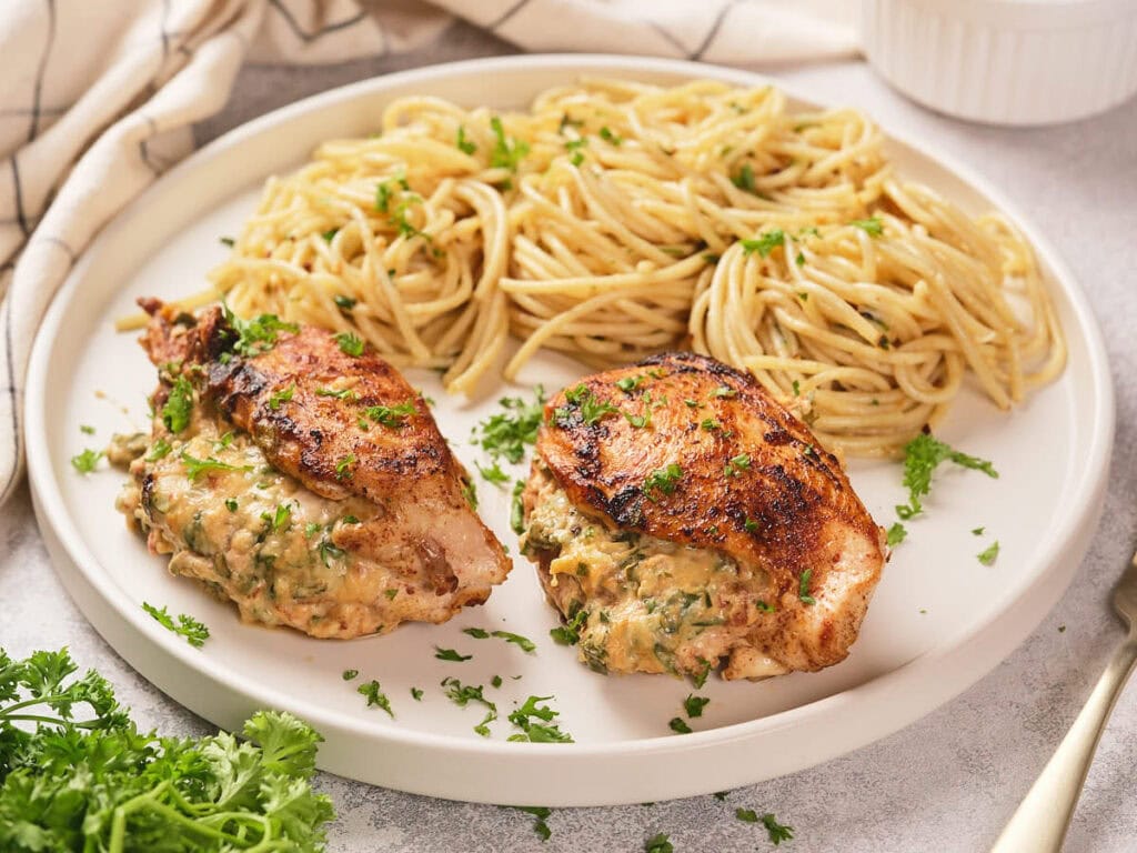 A plate of air fryer stuffed chicken breast and cooked pasta garnished with fresh parsley on a white surface.
