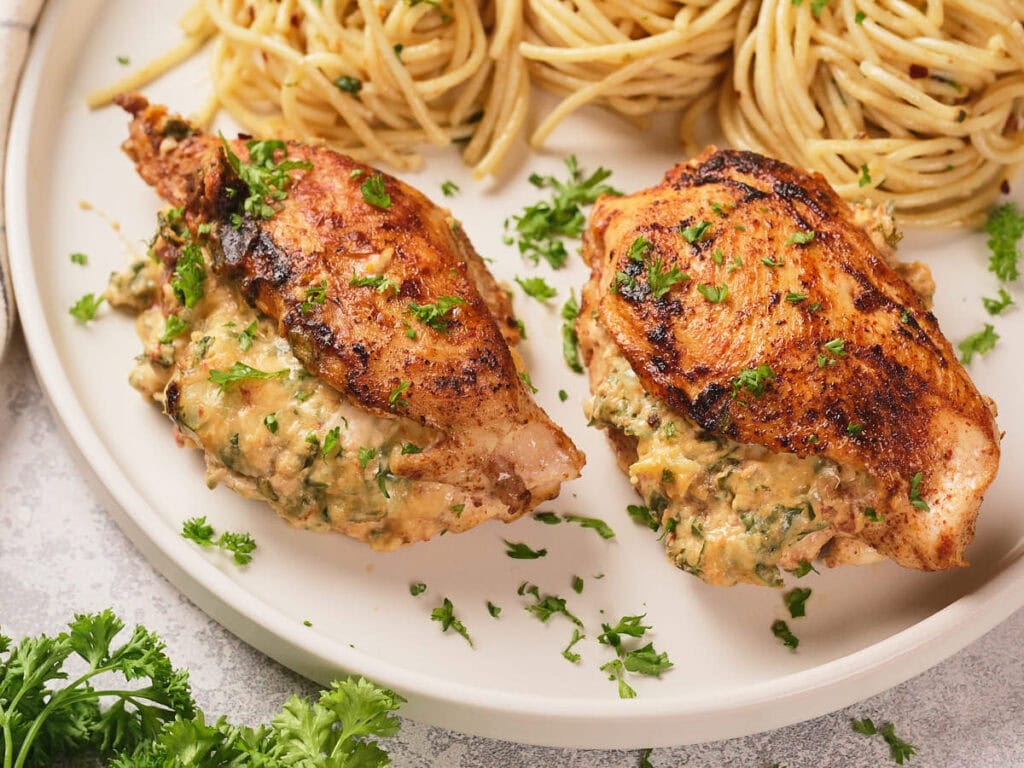 Close-up look of air fryer stuffed chicken breast garnished with fresh parsley on a white plate with cooked pasta next to it.