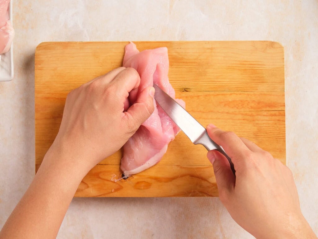 A hand holding a metal knife slicing the chicken breast to form a pocket on a wooden board.
