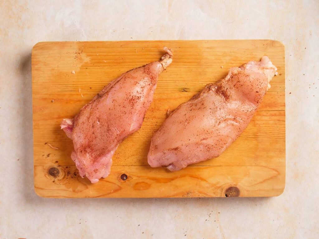 Two chicken breast place on a wooden board seasoned with herbs and spices on a light surface.
