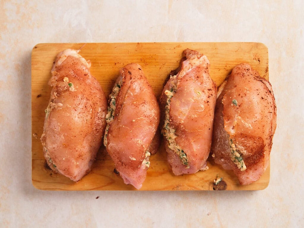Four chicken breasts stuffed with filling on a wooden board seasoned with spices.