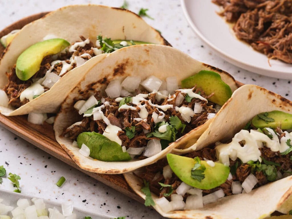 Three barbacoa tacos on a taco holder garnished with white onions, cilantro and avocado.