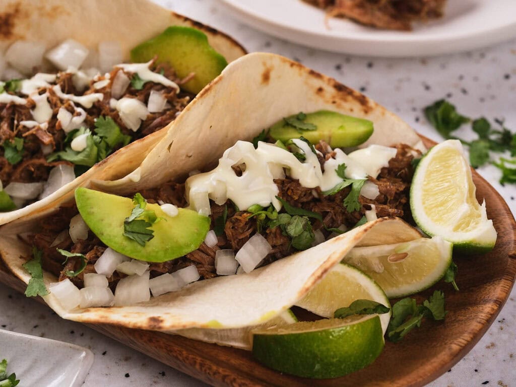 Close-up look of barbacoa tacos with avocado toppings and lime wedges on the side.