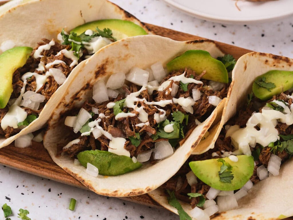 A parallel view of barbacoa tacos garnish with herbs and avocado slices on a wooden taco holder.