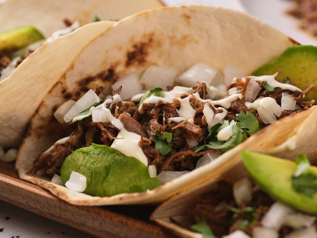 A barbacoa tacos on a wooden taco holder with creamy white sauce, cilantro and a slices of avocados.