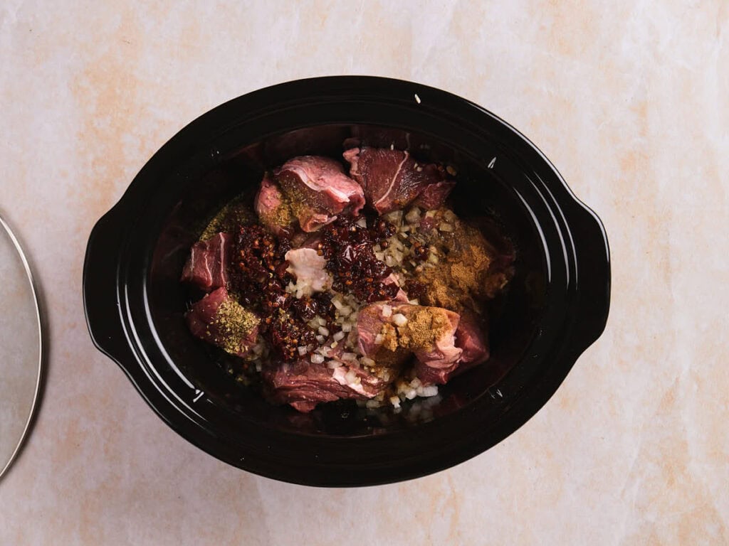 Chunks of raw beef placed inside a black slow cooker with herbs and spices on a light-colored surface.