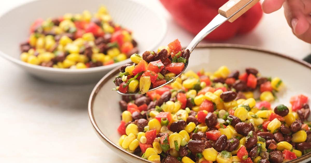 A bowl of black bean salad sits on a table with a spoon and a second small bowl in the background.