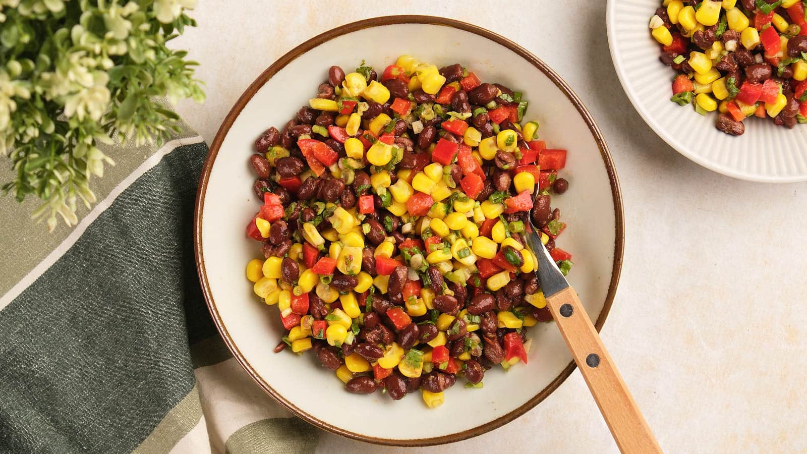 A bowl of black bean salad sits on a table with a spoon and a second small bowl in the background.