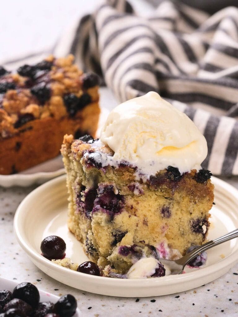 A slice of blueberry buckle topped with a scoop of vanilla ice cream on a plate.