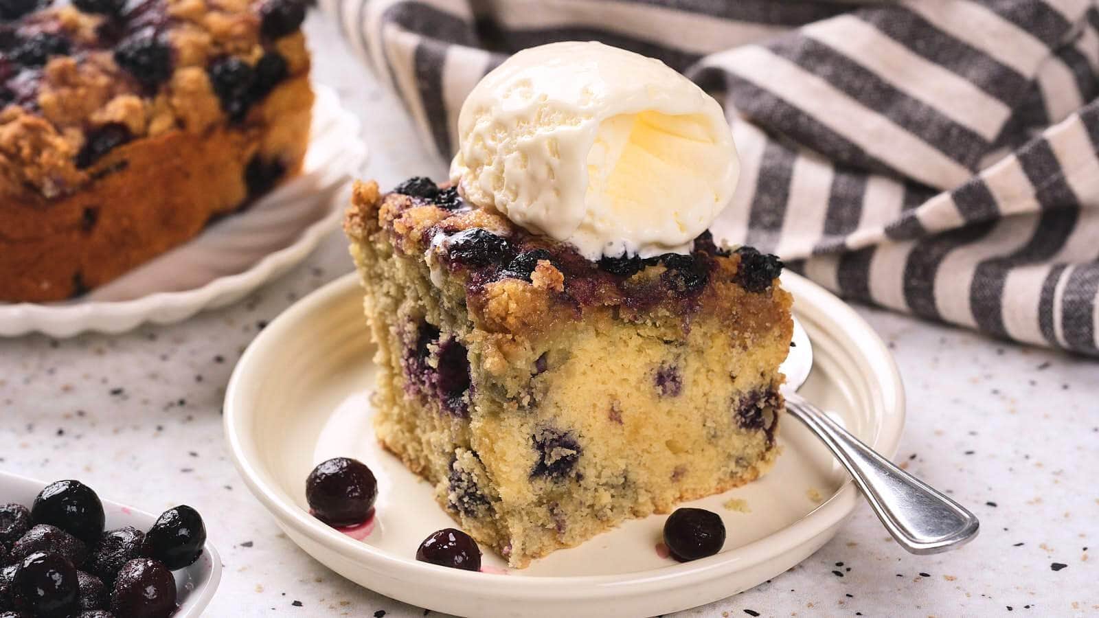 A slice of blueberry buckle topped with a scoop of vanilla ice cream sits on a plate with a spoon, next to a striped cloth.