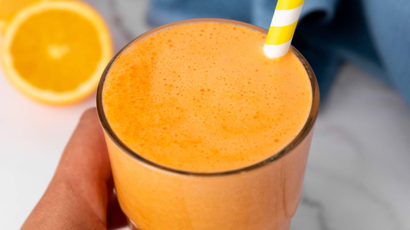 A hand holds a glass of carrot ginger juice with a yellow and white striped straw and a sliced orange is in the background.