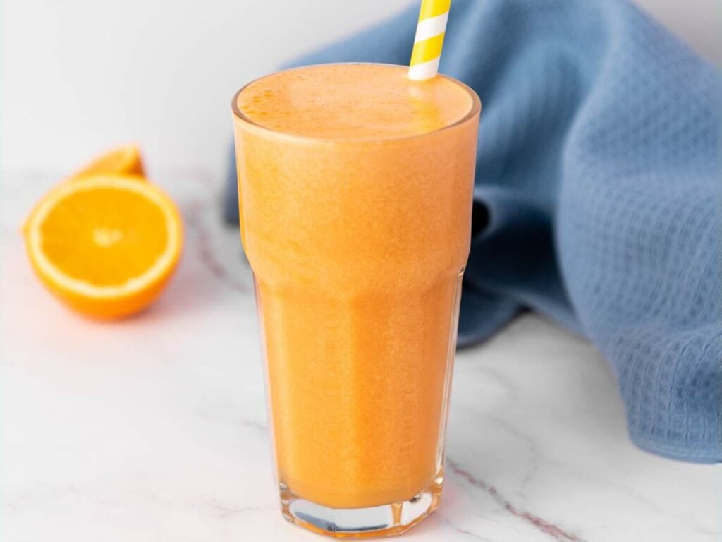A glass of carrot ginger juice with a striped straw sits on a white surface next to a halved orange and a blue cloth.