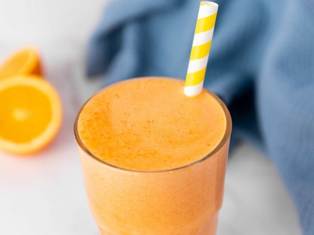 A carrot ginger juice with a yellow and white striped straw, placed on a surface with orange halves and a blue cloth in the background.