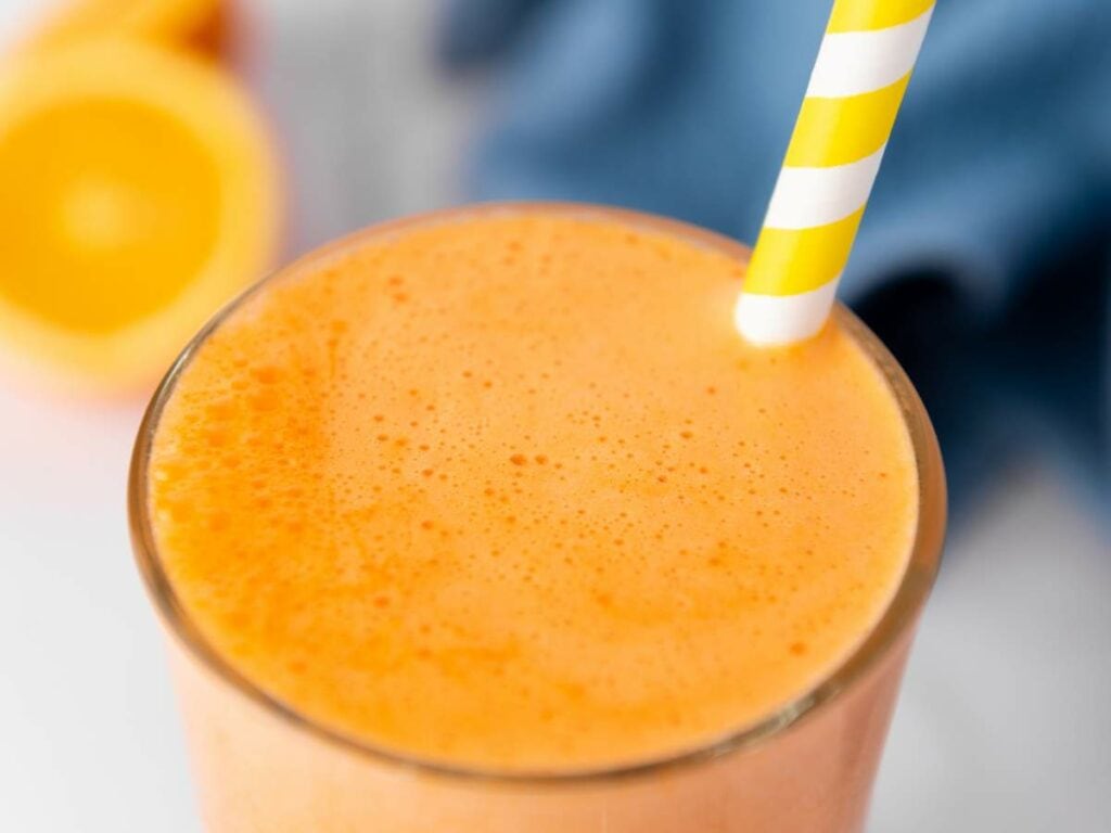 A close-up of a carrot ginger juice in a glass with a yellow-and-white striped straw.