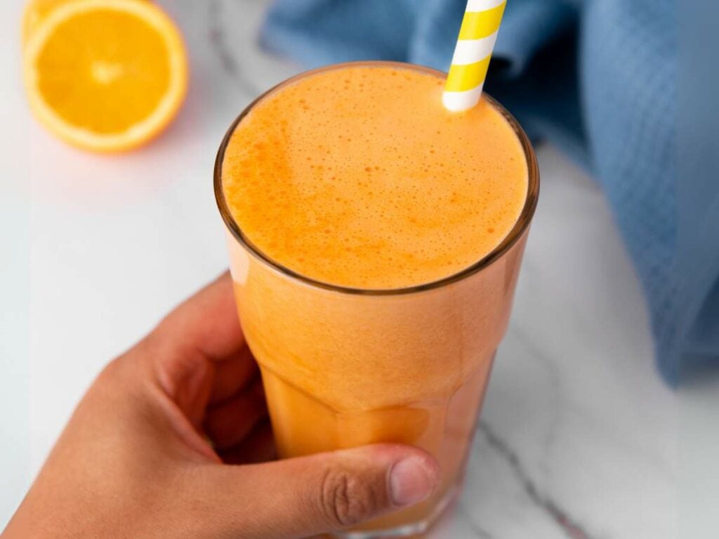 A hand holding a glass of carrot ginger juice with a yellow and white striped straw and half an orange is visible in the background.