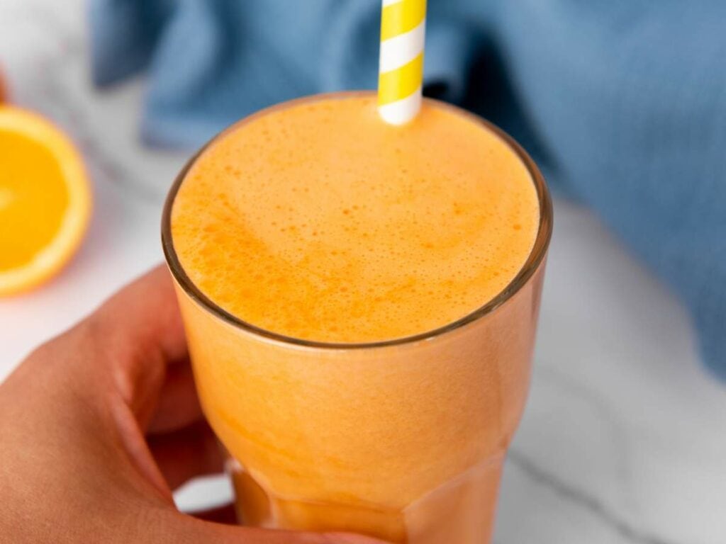 A hand holding a glass of carrot ginger juice with a yellow-and-white striped straw.