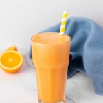 A tall glass of carrot ginger juice with a yellow and white striped straw, placed on a marble surface.