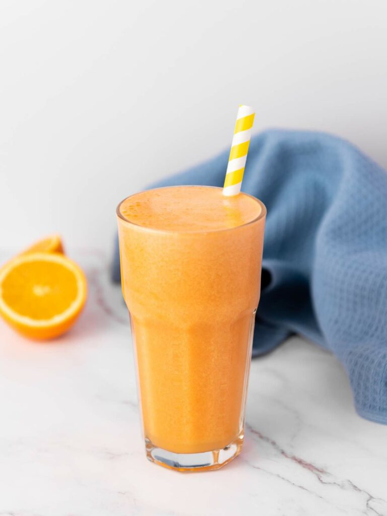 A tall glass of carrot ginger juice with a yellow and white striped straw, placed on a marble surface.