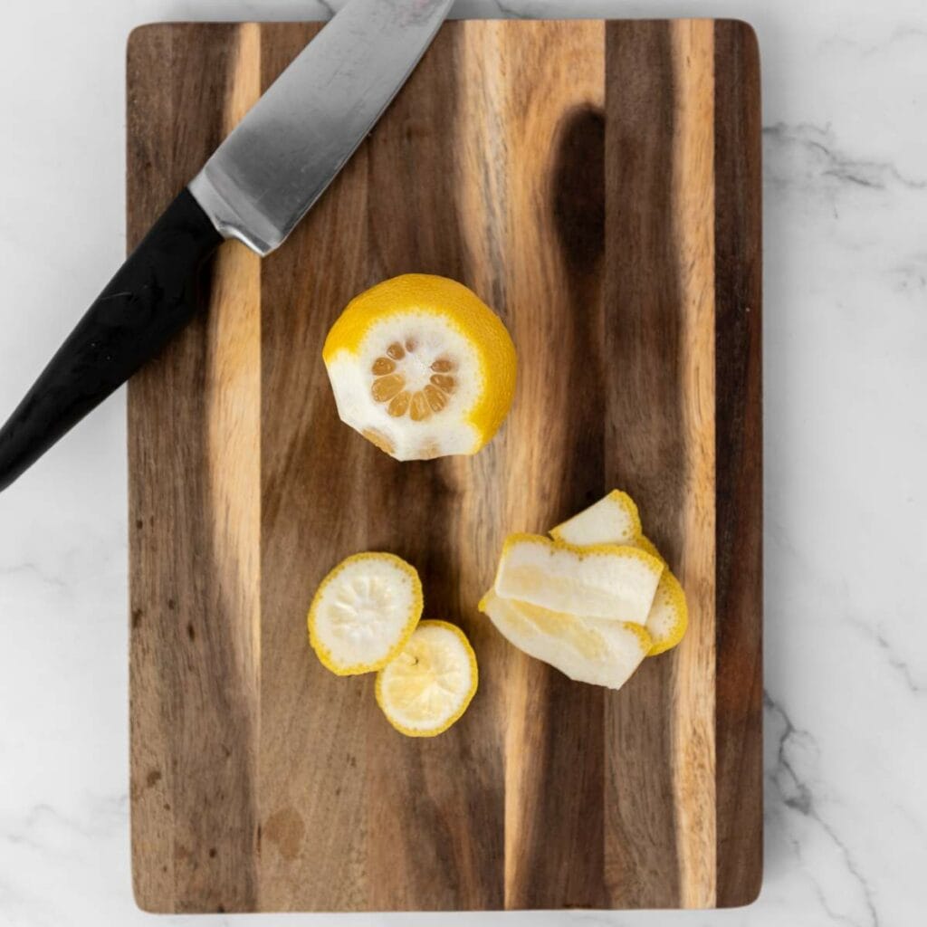 A partially peeled lemon and lemon peel are on a wooden cutting board next to a kitchen knife.