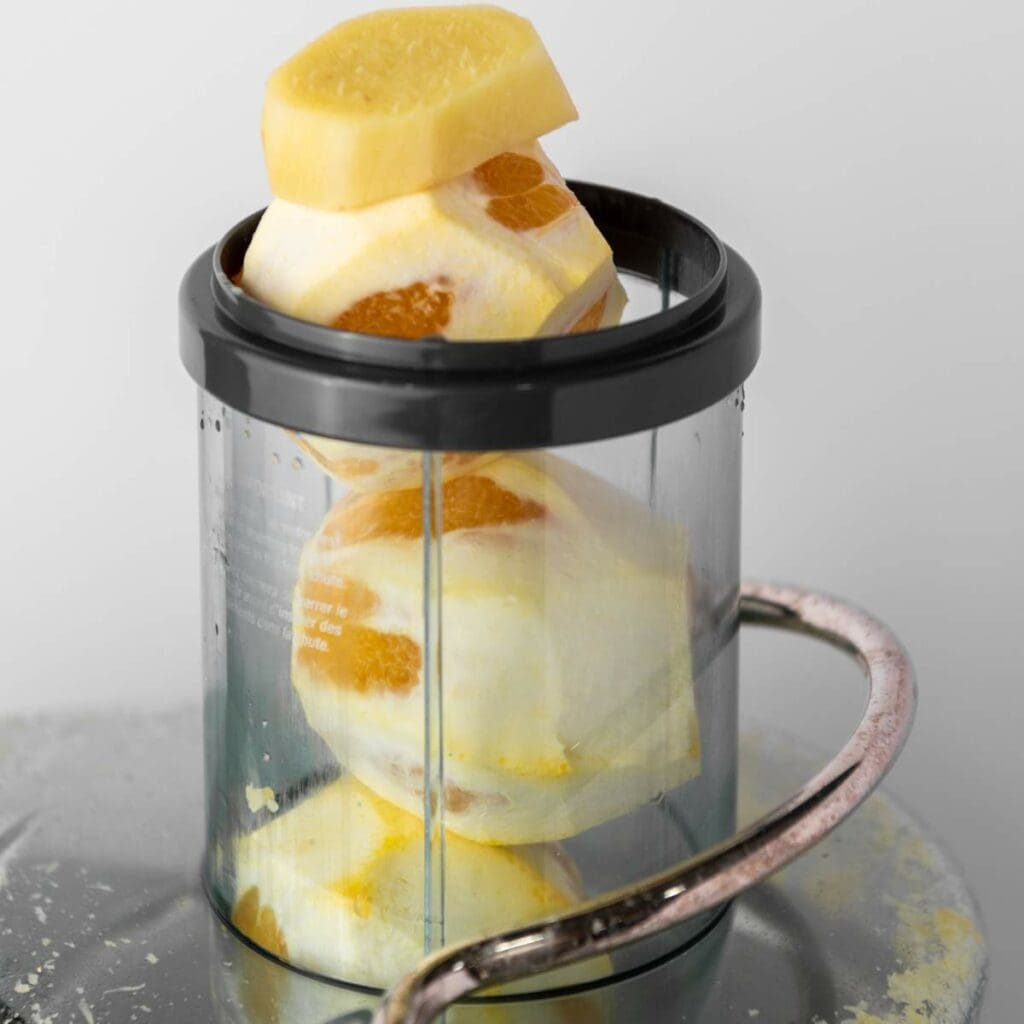 Peeled piece of raw ginger and peeled oranges stacked inside the juicer with a metal handle.