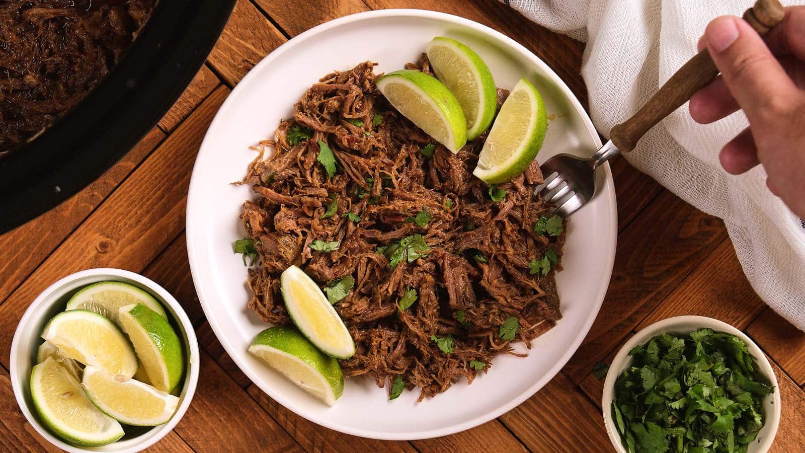 A plate of chipotle barbacoa garnished with cilantro and lime wedges, with a hand holding a fork on a wooden table.