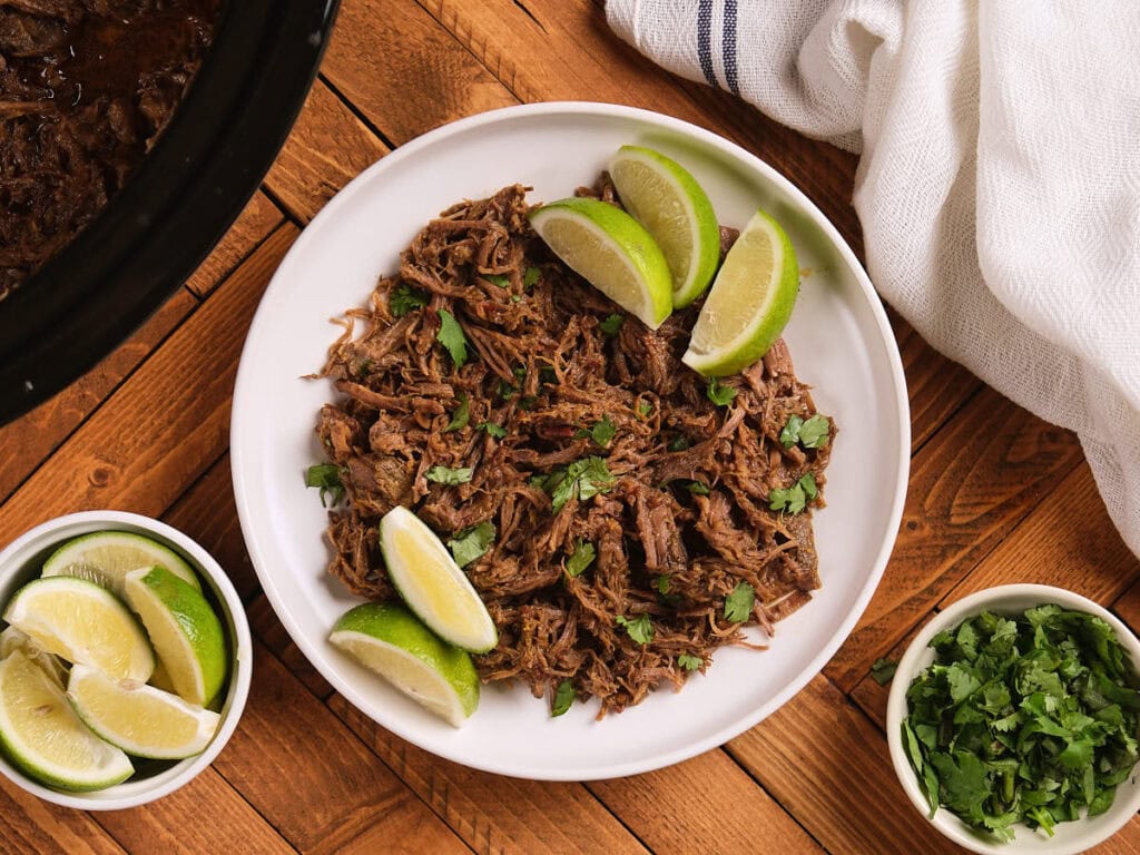 A white plate of chipotle barbacoa garnished with chopped cilantro and lime wedges, on a wooden table next to bowls of lime slices and cilantro.