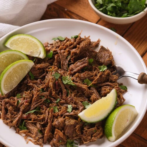 A white plate of barbacoa garnished with chopped cilantro and lime wedges, with a bowl of fresh cilantro nearby on a wooden table.