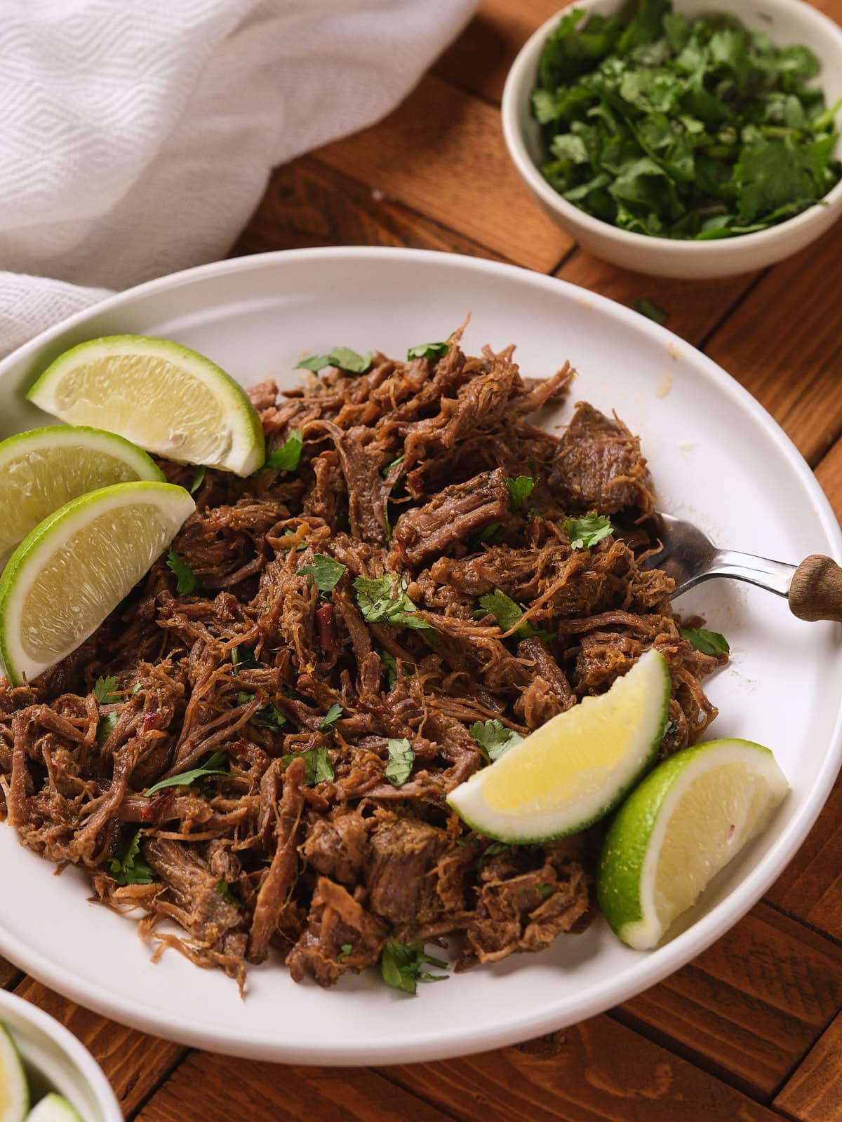A white plate of barbacoa garnished with chopped cilantro and lime wedges, with a bowl of fresh cilantro nearby on a wooden table.