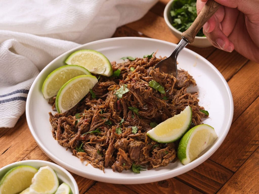 Chipotle barbacoa garnished with lime wedges and chopped herbs, with a hand using a fork, on a wooden table.