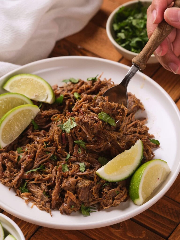A chipotle barbacoa garnished with a hand using a fork to pick up some meat with bowl of chopped herbs is in the background.