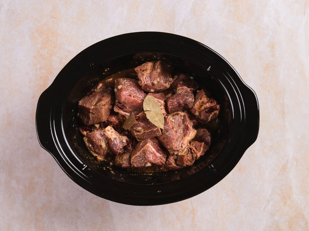 A black slow cooker filled with raw chunks of beef and bay leaves, placed on a light-colored countertop.