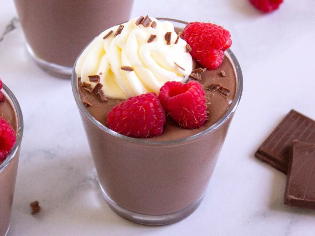 A glass of chocolate cottage cheese pudding and chocolate shavings sits on a white surface next to pieces of chocolate.