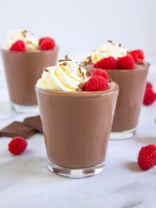 Three glass of chocolate cottage cheese pudding topped with whipped cream, chocolate shavings, and fresh raspberries.