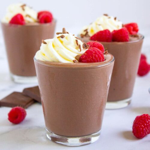 Three glass of chocolate cottage cheese pudding topped with whipped cream, chocolate shavings, and fresh raspberries.