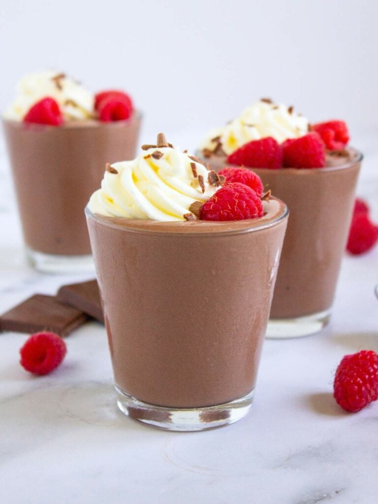 Three glass of chocolate cottage cheese pudding topped with whipped cream, chocolate shavings, and fresh raspberries.