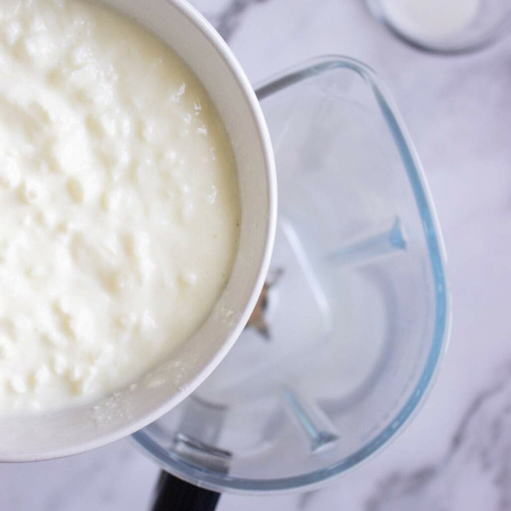 A bowl of cottage cheese is being poured into a blender on a marble countertop.