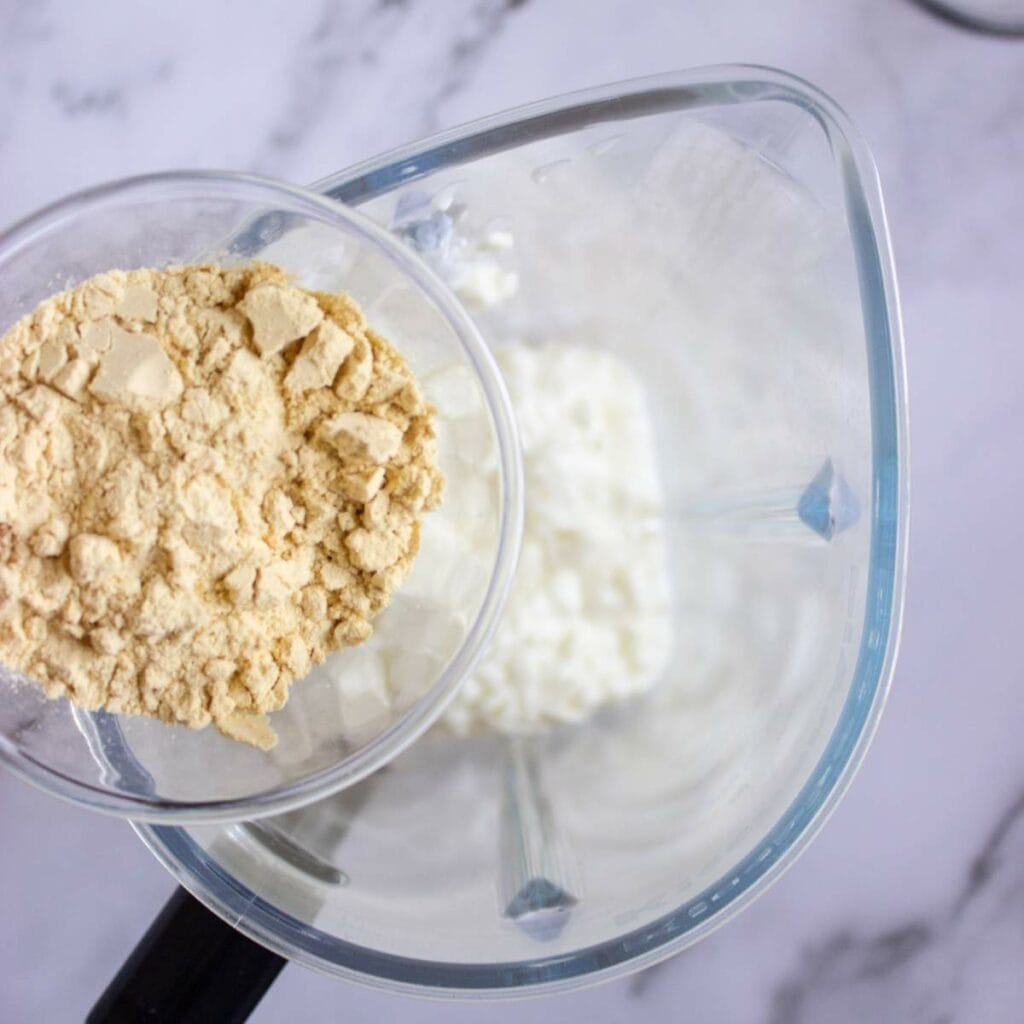 A measuring cup of protein powder is being poured into a blender.