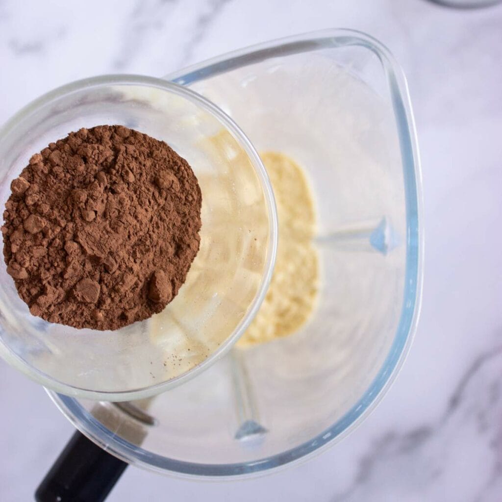 A glass cup filled with cocoa powder is being held above a blender.