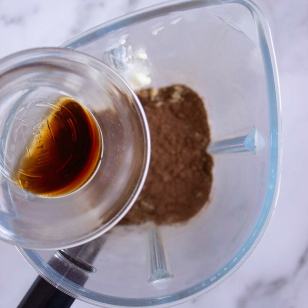 A glass blender contains cocoa powder and a small glass cup of vanilla extract.