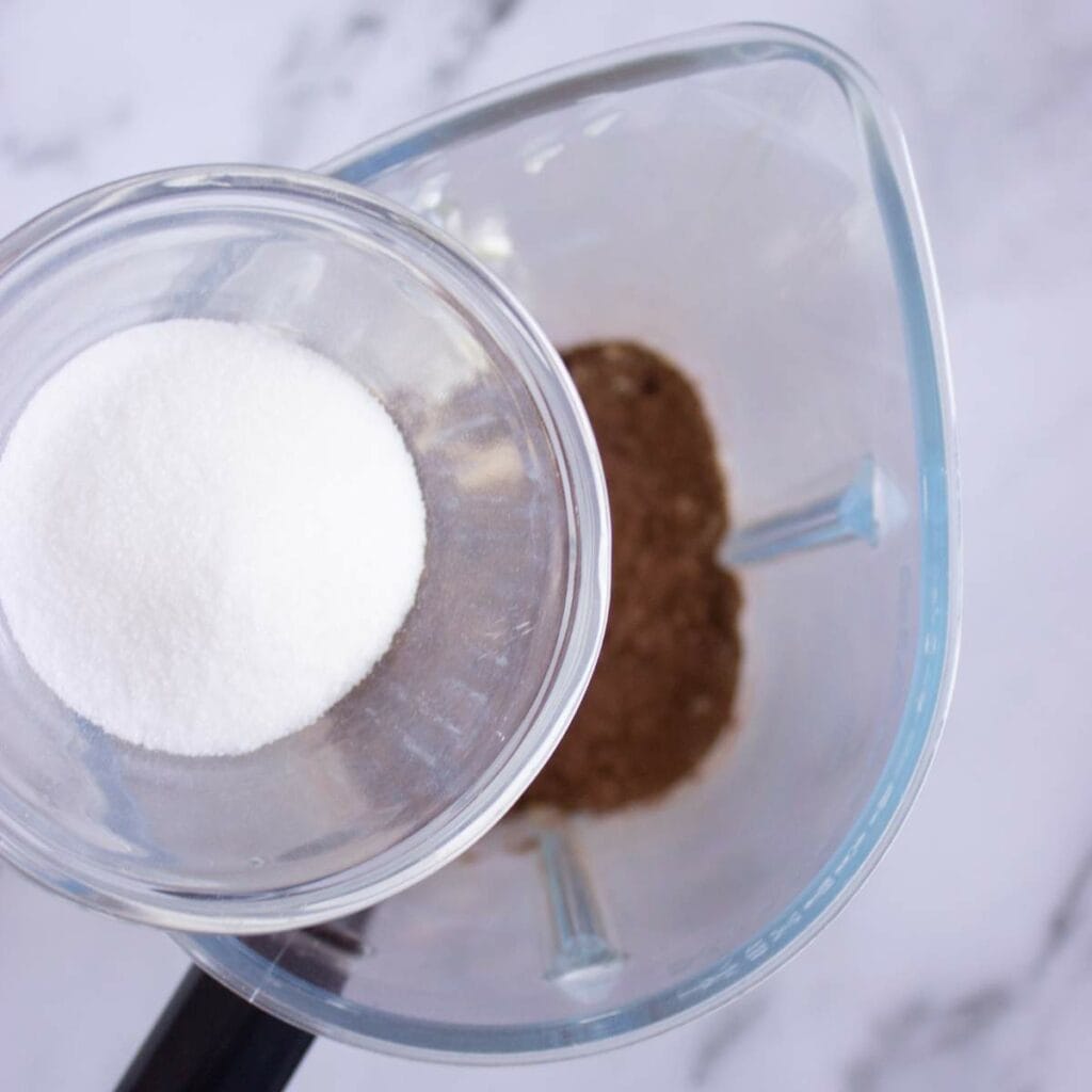 A glass bowl of granulated sugar is being poured into a blender.