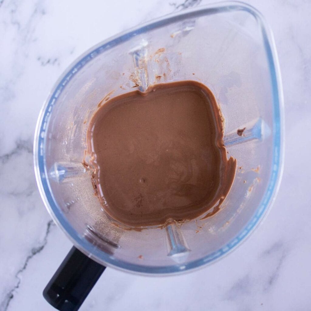 A blender containing a thick, light brown liquid mixture on a marble surface.