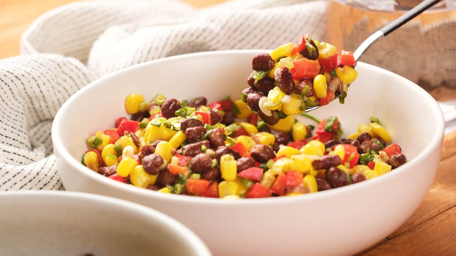 A bowl of corn and black bean salad containing black beans, corn, diced red bell pepper, and herbs, with a spoon lifting a portion.