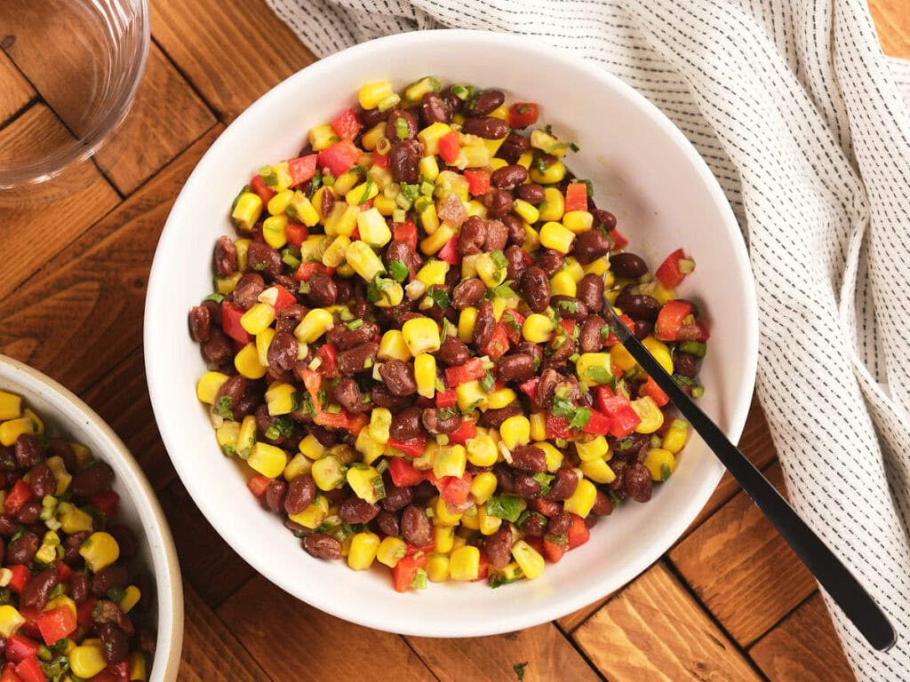 A white bowl filled with corn and black bean salad with a metal spoon, on a wooden table next to a white cloth.